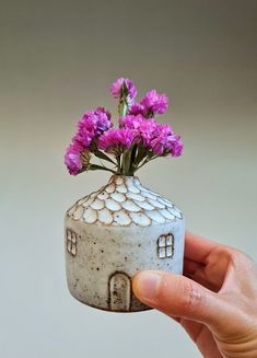 a hand holding a small white vase with purple flowers in it and a tiny house on the inside