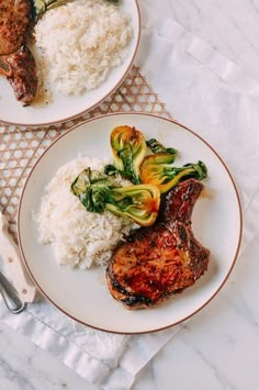 two plates with meat, rice and vegetables next to silverware on a white table