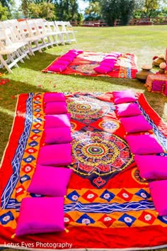 an outdoor ceremony set up with bright pink and orange blankets, white chairs and flowers on the grass