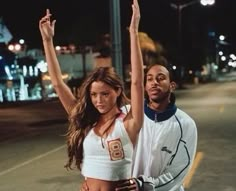 a man and woman standing next to each other on the street at night with their arms in the air