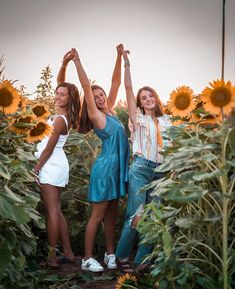 three young women standing in a sunflower field with their arms up and hands raised