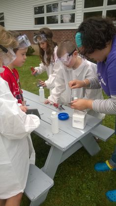 several children in white lab coats and goggles around a picnic table with test tubes on it