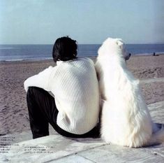 a man sitting on the beach next to two large white dogs, both looking out at the ocean