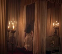 a woman standing in front of curtains with candles on either side of her head and looking out the window