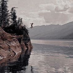 a person jumping into the water from a cliff on a lake with mountains in the background