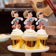 three cupcakes with cartoon characters on them sitting on a cake stand in front of other cupcakes