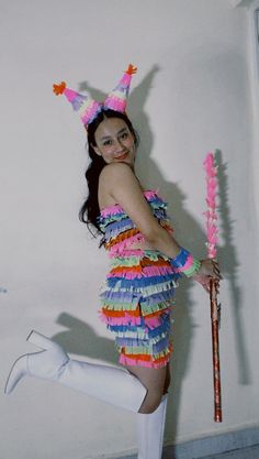 a woman in a colorful dress holding a stick and wearing a party hat while standing on one leg