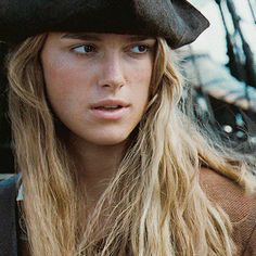 a woman with long blonde hair wearing a black hat and looking at the camera while standing in front of a ship