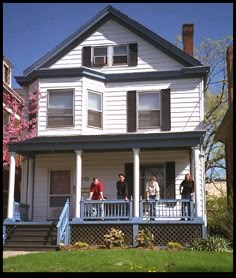 three people standing on the porch of a white two - story house with blue trim