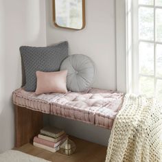 a pink bench with pillows on it next to a mirror and bookcase in front of a window