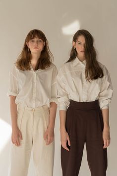 two women standing next to each other wearing white shirts and brown pants, both with their hands in their pockets