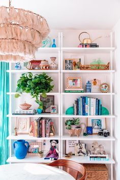 a white book shelf filled with books next to a table and blue curtained window