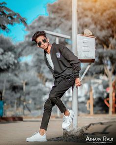 a young man leaning against a post wearing sunglasses and a black suit with white sneakers