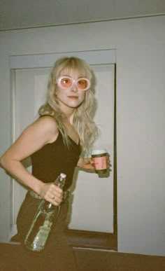 a woman with pink glasses holding a can and a drink in her hand while standing next to a mirror