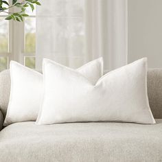 two white pillows sitting on top of a couch next to a potted plant in front of a window