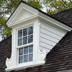 a white window on the side of a house with black shingles and trees in the background