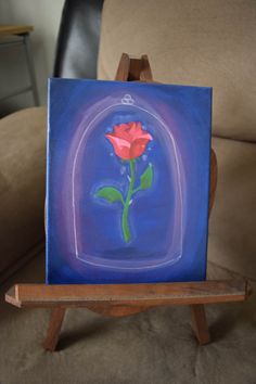 a painting of a red rose in a glass case on a wooden easel with a blue background