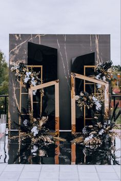 a black and gold wedding backdrop with flowers on the front door, surrounded by greenery