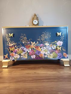 a blue dresser with flowers painted on the front and gold trimmings, along with a clock