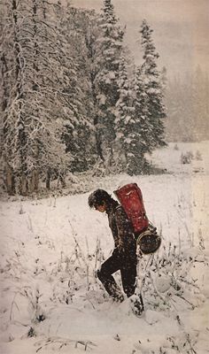 a man walking through the snow carrying a red backpack