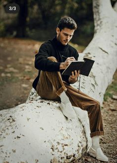 a man sitting on top of a white log reading a book next to a tree