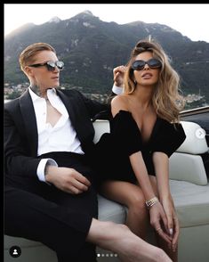 a man and woman sitting on the back of a boat wearing sun glasses, one in a black dress