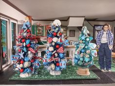 a woman standing next to three christmas trees in a living room with blue and red decorations