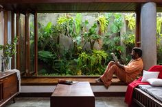 a man sitting on a bed in front of a window reading a book while surrounded by greenery