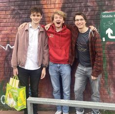 three young men standing in front of a brick wall with graffiti on it and holding shopping bags