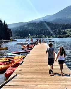 two people walking down a dock towards boats on the water and mountains in the background