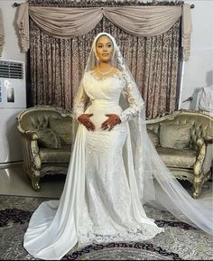 a woman in a white wedding gown and veil standing next to a couch with curtains