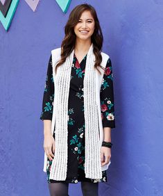 a woman standing in front of a blue wall wearing a white vest and black dress