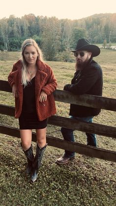 a man and woman standing next to each other near a wooden fence in a field