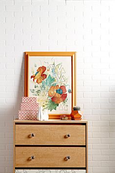 a wooden dresser topped with drawers next to a white brick wall and framed artwork on the wall