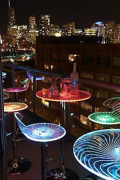 illuminated chairs and tables on a balcony overlooking the city skyline at night, lit up with colorful lights