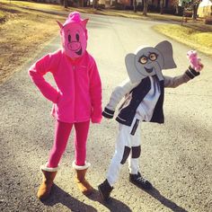 two people in costumes standing on the street with one holding up a paper elephant and the other pointing at something