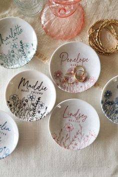 six personalized wedding rings are displayed in small bowls on a white tablecloth with floral designs