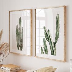 two framed cactus prints on a wall next to a table with books and vases