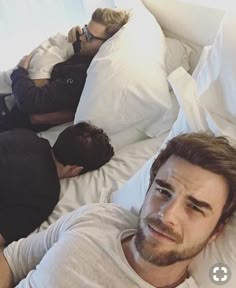 two men laying in bed and one is looking at the camera while another man sleeps