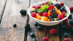 a white bowl filled with fresh fruit on top of a wooden table next to blueberries, raspberries and peaches