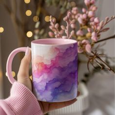 a person holding a coffee mug with pink and purple paint on it in front of some flowers