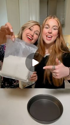 two women standing next to each other in front of a pan on a counter top