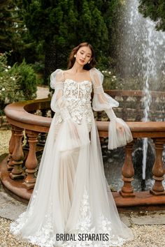 a woman standing in front of a fountain wearing a wedding dress with long sleeves and sheer tulle