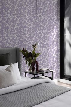 a bed sitting next to a window with purple wallpaper on it and a vase filled with flowers