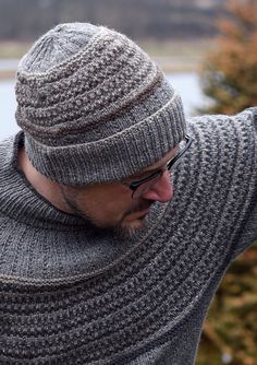 a man wearing glasses and a knitted hat looks at his cell phone while standing in front of a lake