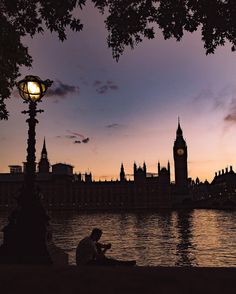 a person sitting on a bench next to the water at dusk with a clock tower in the background