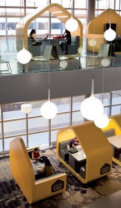 people are sitting at desks in an office with yellow and gray furniture, large windows, and hanging lights