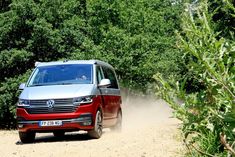 a red van driving down a dirt road next to green bushes and trees on a sunny day