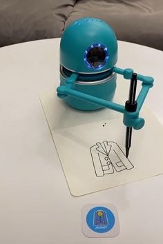 a blue robot sitting on top of a white table next to a pen and paper