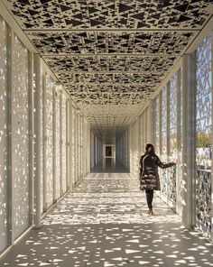 a woman walking down a long hallway with lots of windows on both sides and shadows from the ceiling